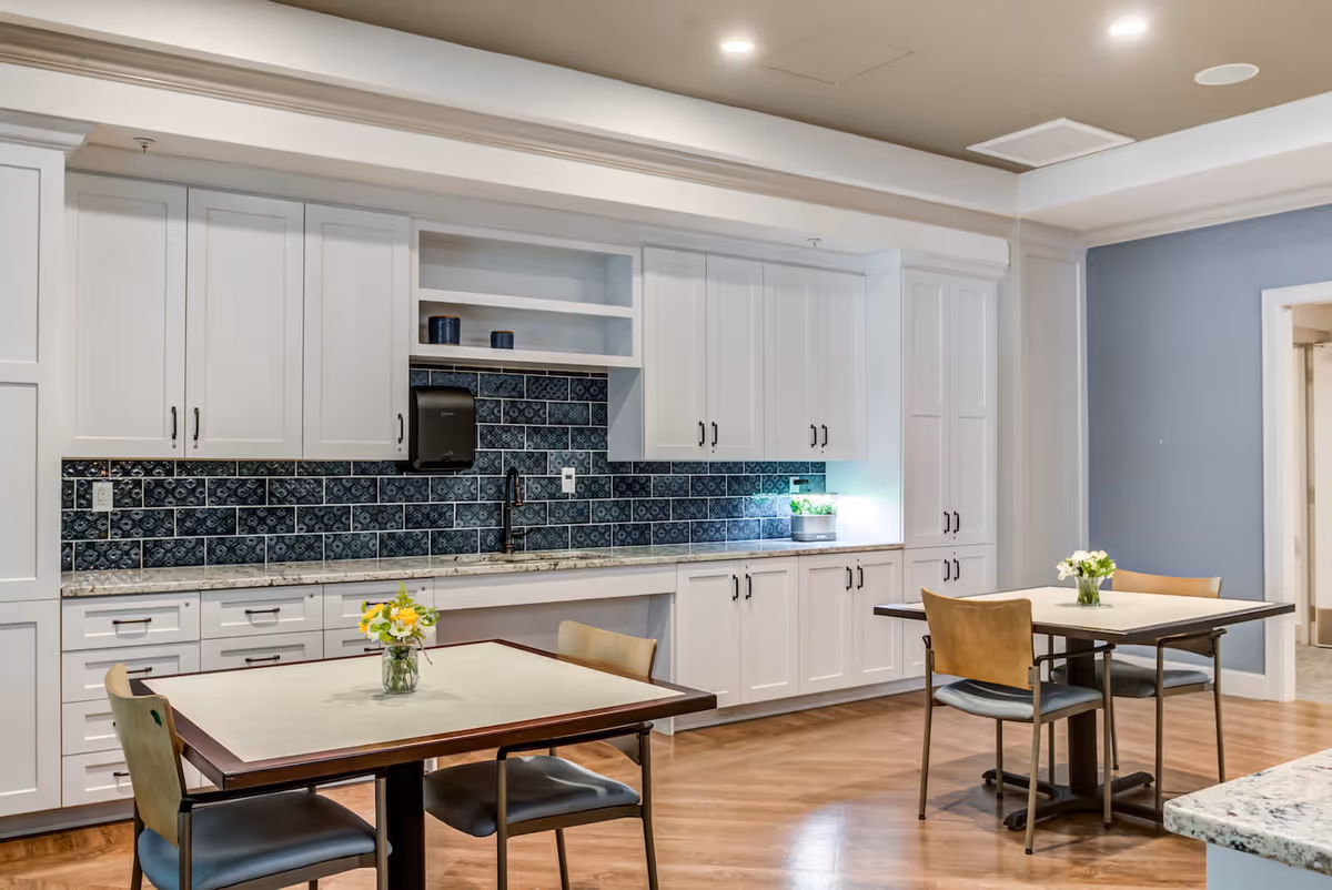 A bright and clean dining area with two square tables, each with two chairs. The tables have small vases with flowers. Behind the tables is a wall with white cabinetry and a countertop, featuring a dark blue patterned tile backsplash. The floor is wooden, and the walls are painted light blue and white.