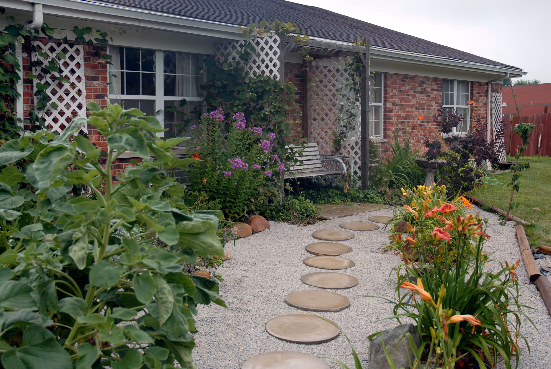 Front of a single-story brick building with a gravel path of circular stepping stones leading through flower beds to a white trellis and bench by the windows.