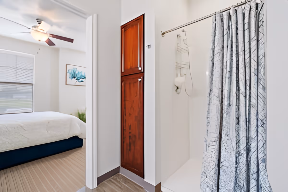 View of a bathroom with a shower and a curtain on the right side, and a glimpse into a bedroom with a bed, ceiling fan, window with blinds, and wall art on the left side. There is a wooden cabinet between the bathroom and bedroom.