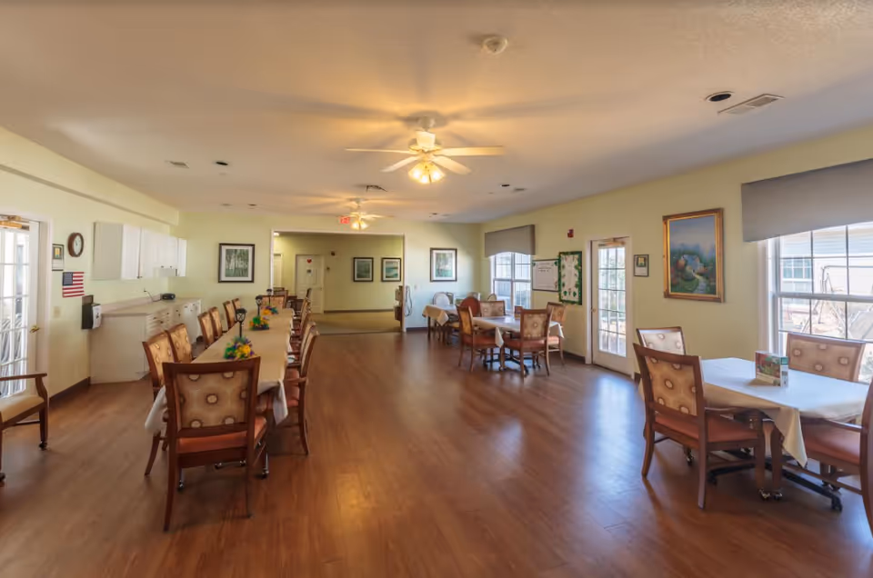 A spacious dining room with several tables covered with white tablecloths and surrounded by wooden chairs with patterned cushions. The room has wooden flooring, light yellow walls, ceiling fans with lights, and large windows with gray blinds letting in natural light. There are framed pictures on the walls and a door leading outside.