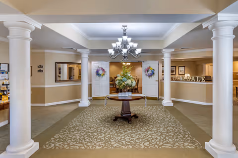 Well-lit lobby with white columns, a central table holding a floral arrangement under a chandelier, and a reception desk in the background.