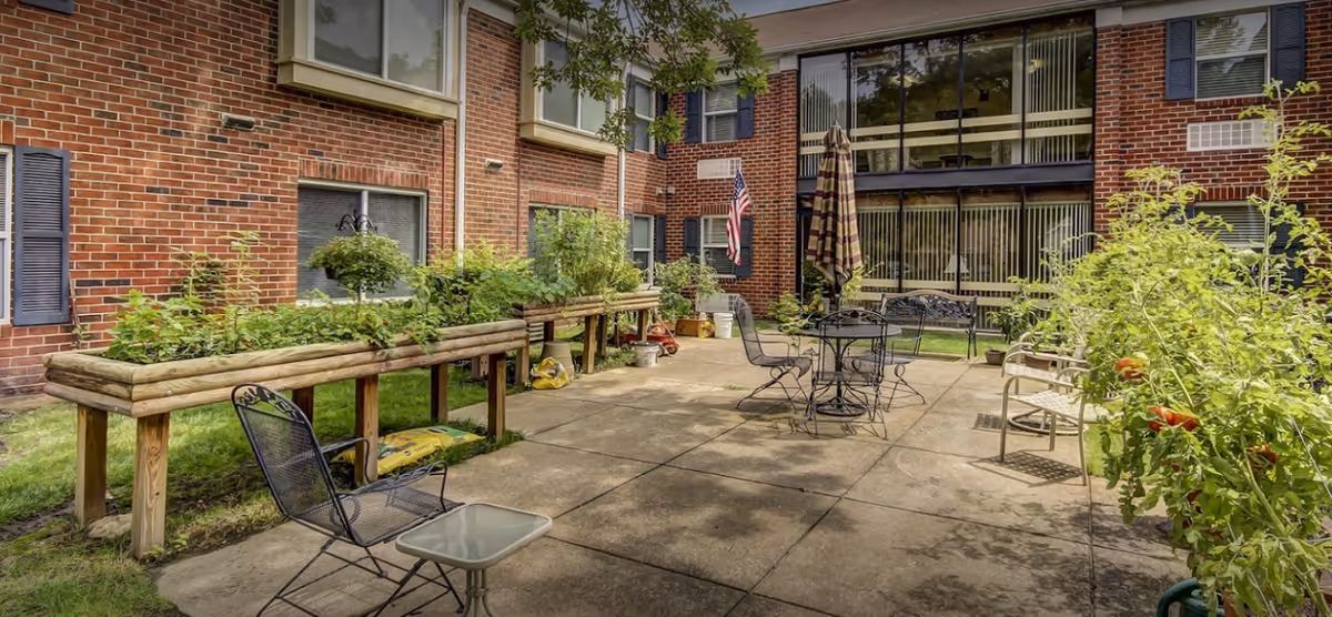Sunlit courtyard with raised wooden planters, patio tables and chairs, an umbrella and an American flag in front of a brick senior living building.