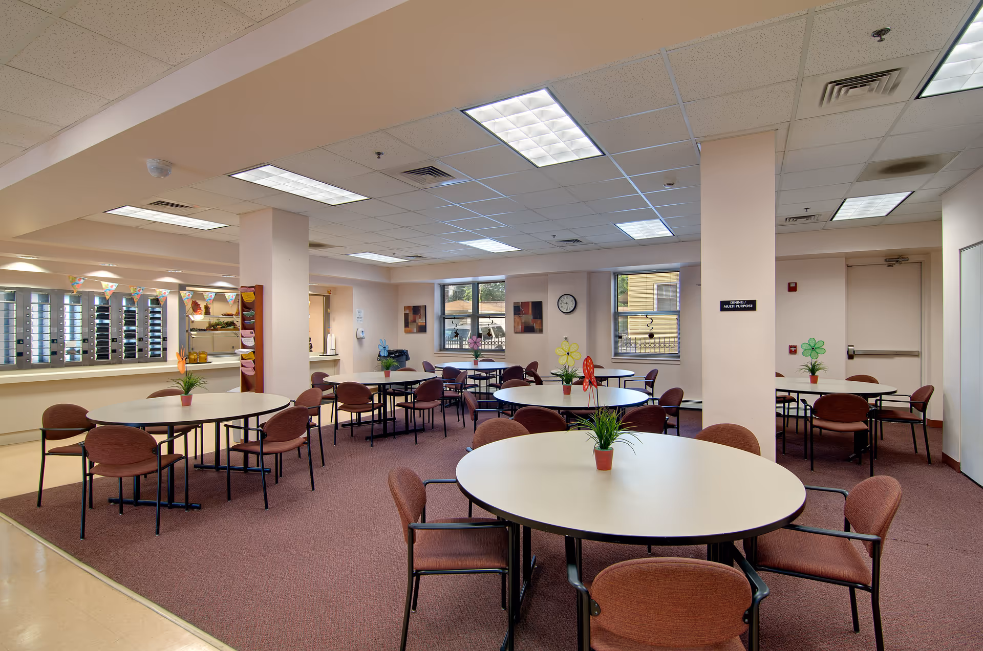 Communal dining/activity room with multiple round tables and chairs, potted plants on the tables, and a service counter along the left wall.