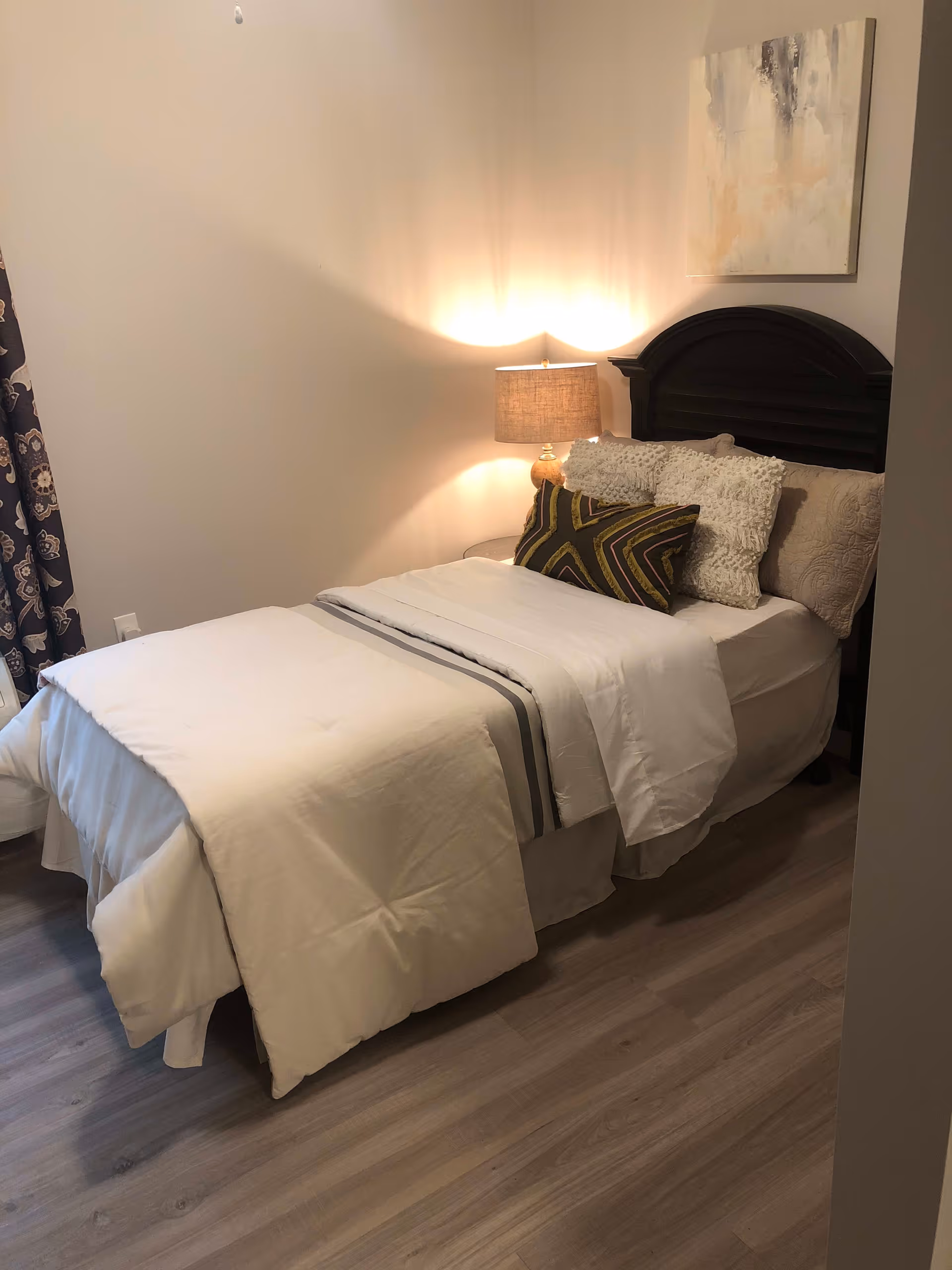 A neatly made single bed with white bedding and decorative pillows in a room with light-colored walls and wood flooring. A bedside table with a lamp is next to the bed, and a piece of abstract art hangs on the wall above the headboard.