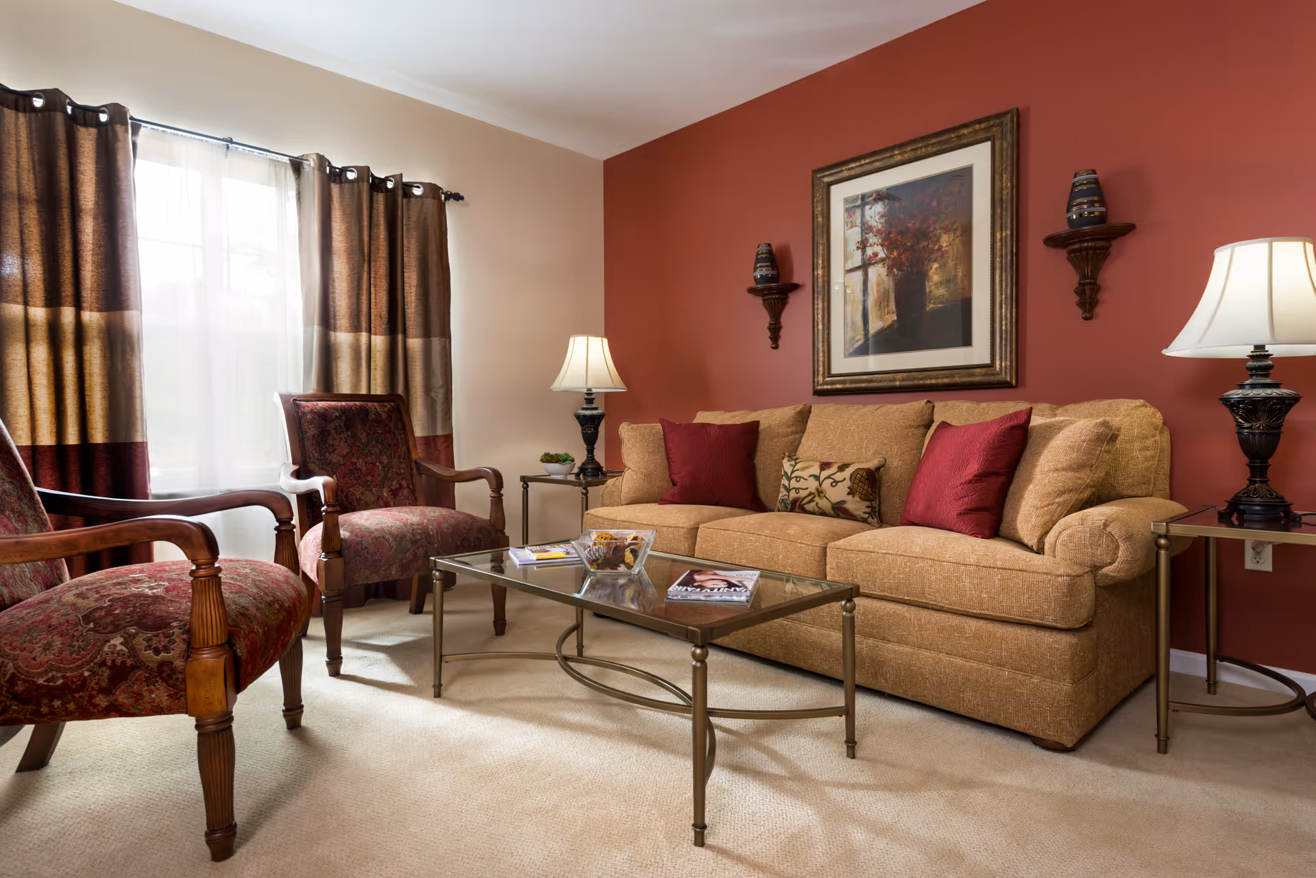 A cozy living room with a beige sofa adorned with red and patterned cushions, two upholstered armchairs with wooden frames, a glass coffee table with magazines and decorative items, two side tables each with a lamp, and a large window with brown and beige curtains. The walls are painted beige and red, with a framed artwork and two decorative wall shelves.