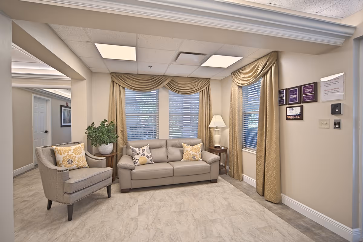 A cozy living room area in a senior living facility with a beige leather sofa adorned with decorative pillows, a matching armchair with a floral pillow, a side table with a potted plant, and another side table with a lamp. The room has large windows with gold curtains and blinds, beige walls, and a light-colored floor. Several plaques and a keypad are mounted on the wall to the right.