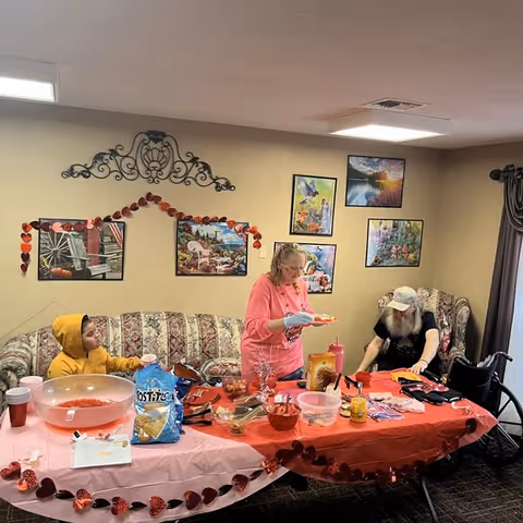 Three people gathered around a long table covered with red decorations and snacks in a cozy decorated common room.