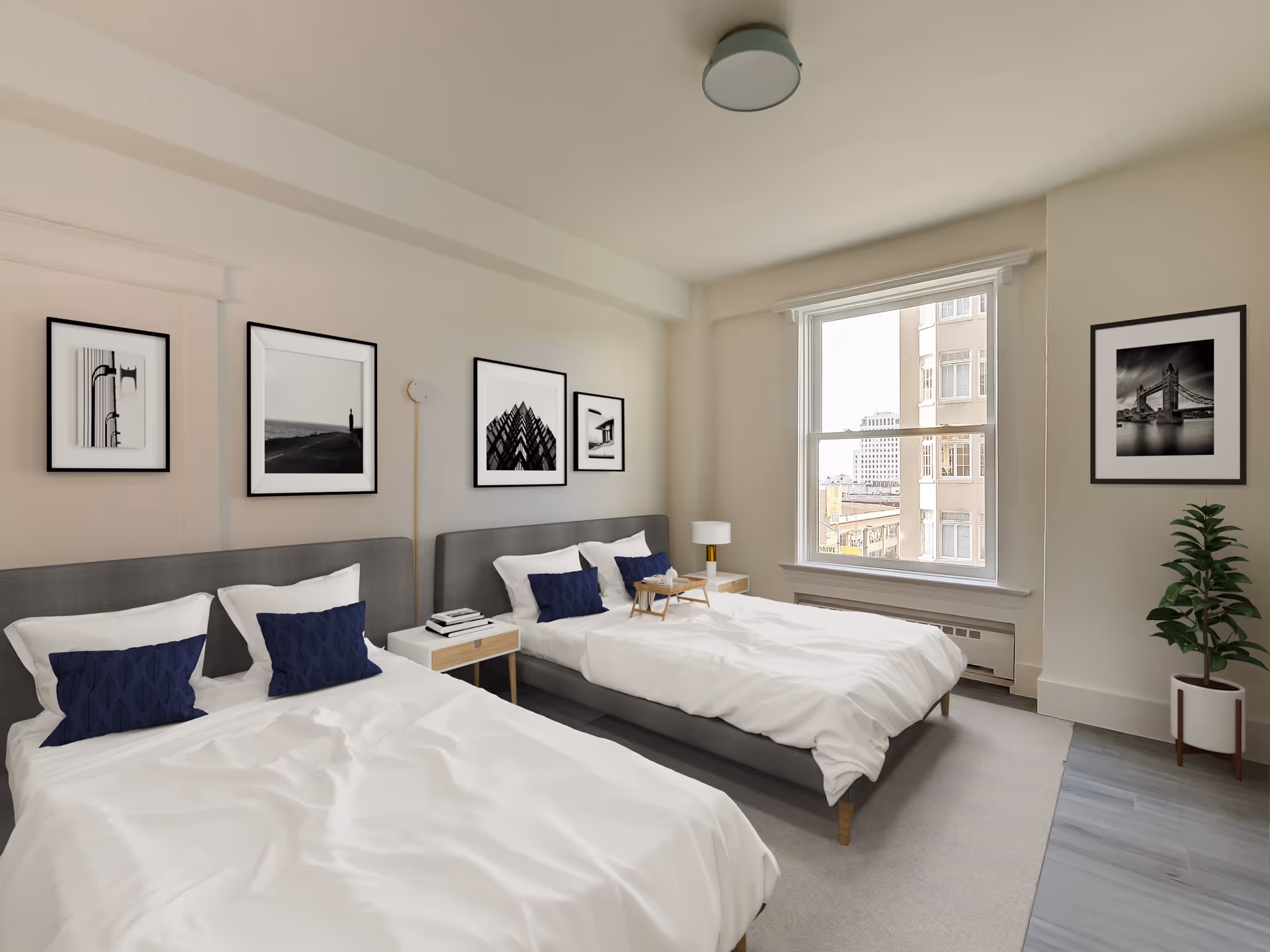 A bright bedroom with two double beds featuring gray upholstered headboards, white bedding, and dark blue accent pillows. Between the beds is a small wooden nightstand with books and a lamp. The walls are decorated with black and white framed photographs. A large window lets in natural light and shows a cityscape outside. A potted plant is placed in the corner near the window.