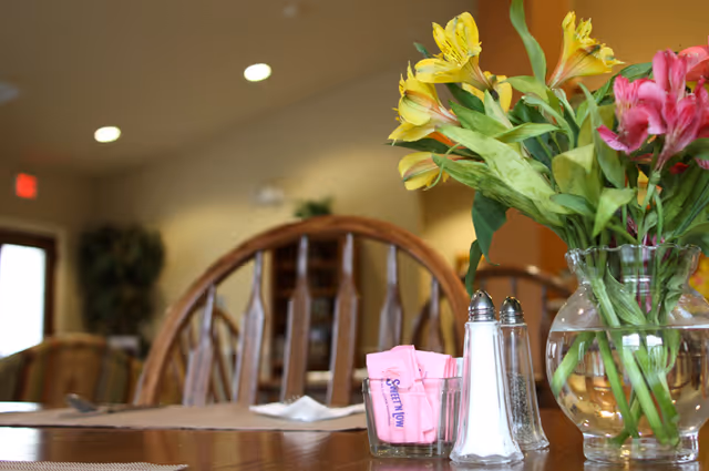 Close-up of a dining table with a glass vase holding yellow and pink flowers, a glass container with pink Sweet'N Low packets, and salt and pepper shakers. Wooden chairs and a softly lit room are visible in the background.