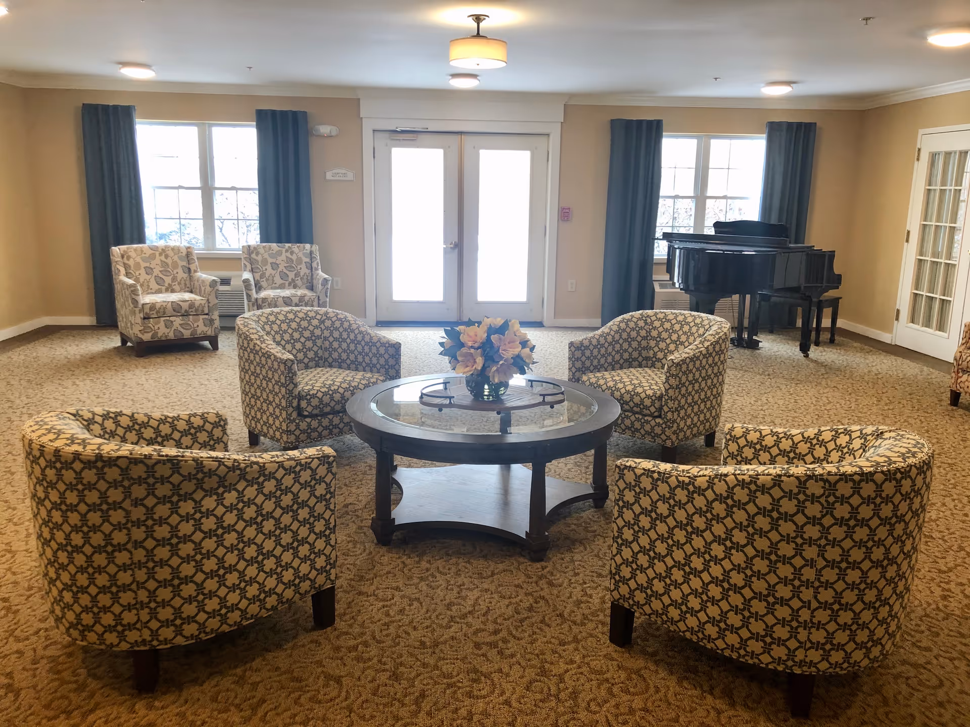 A spacious senior living common area with four patterned armchairs arranged around a round glass-top coffee table with a flower arrangement. In the background, there are two floral armchairs near windows with dark curtains, a black grand piano, and double glass doors letting in natural light. The room has beige walls and carpeted flooring.