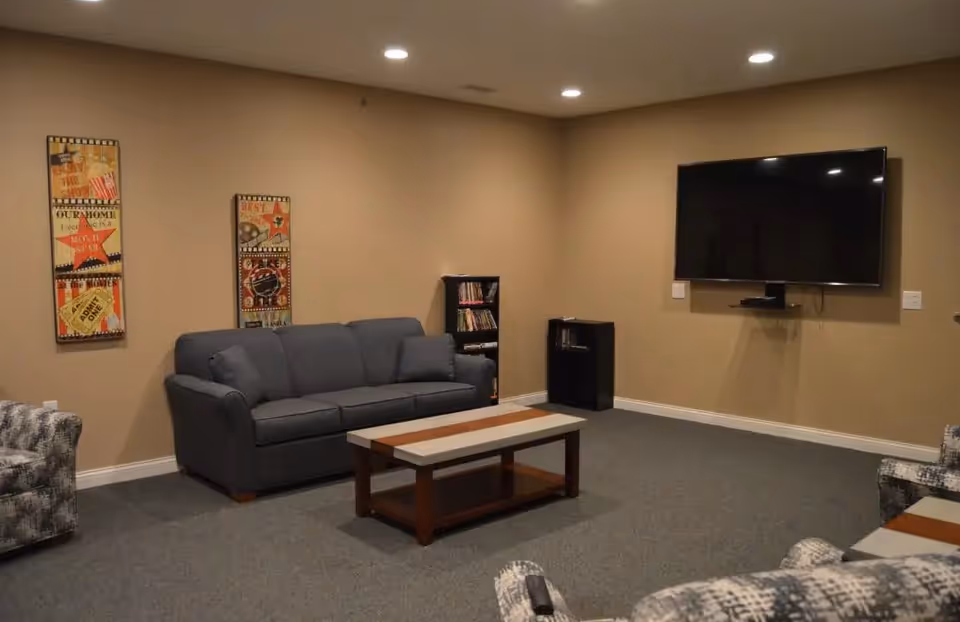 A cozy living room with a gray sofa, patterned armchairs, a wooden coffee table, two small bookshelves, and a large flat-screen TV mounted on a beige wall. The room has carpeted flooring and two decorative wall hangings.