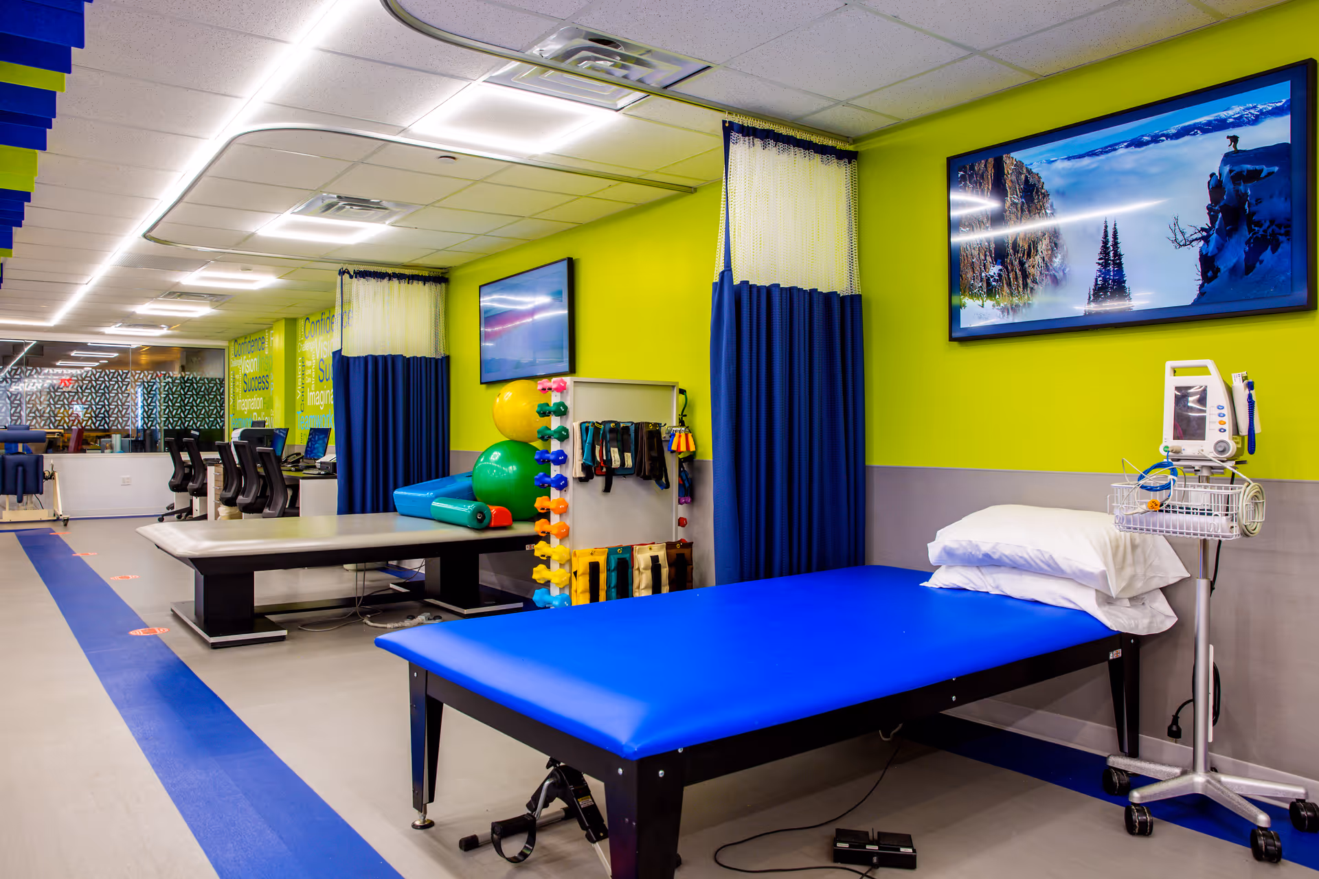 Bright rehabilitation room with blue therapy tables, exercise equipment, and a wall-mounted TV.