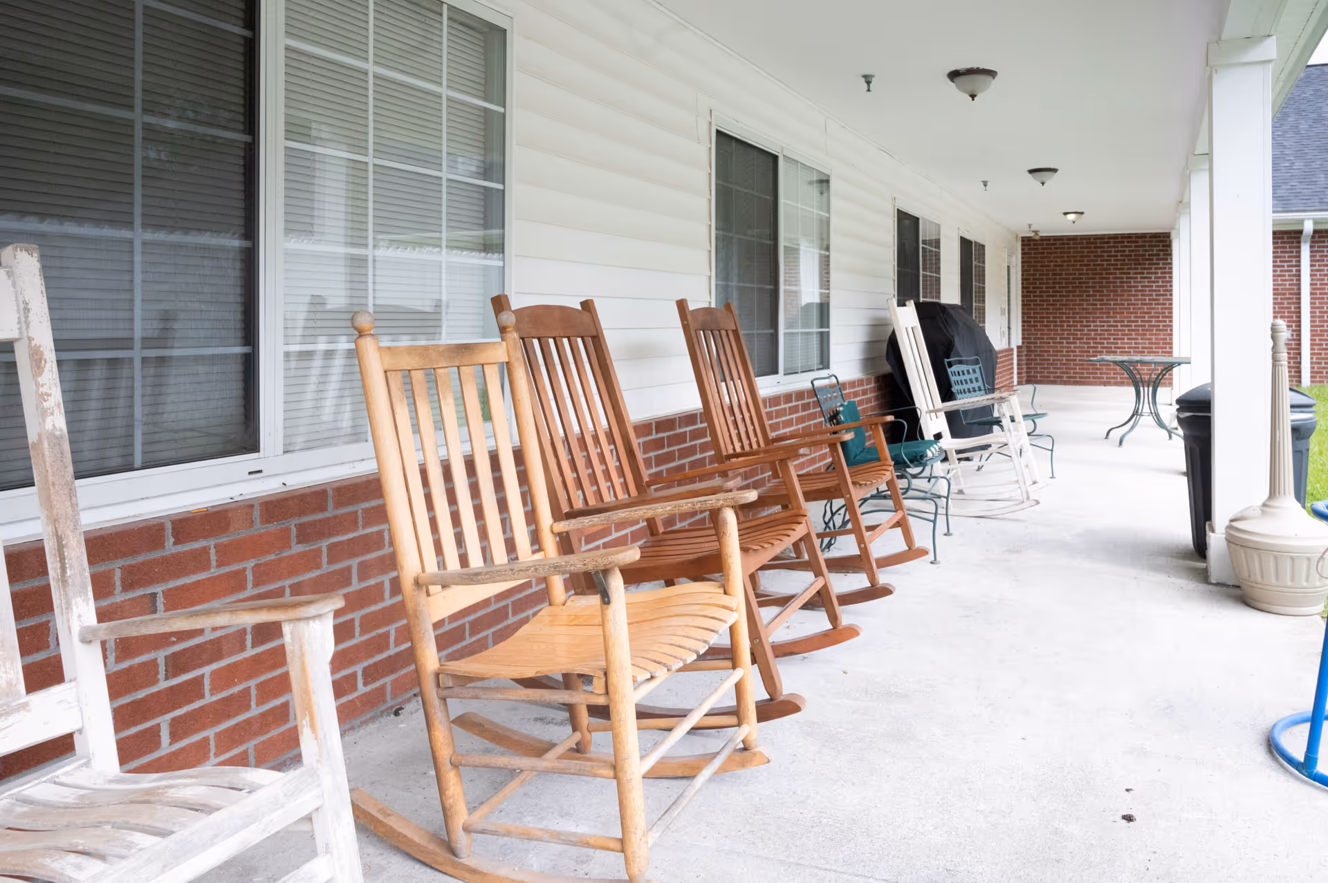 A covered outdoor porch area with several wooden rocking chairs and metal chairs lined up against a brick and white siding wall with windows. There is a small round table and a grill further down the porch.
