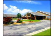 Exterior view of a single-story assisted living facility building with a covered entrance, surrounded by landscaped greenery and a driveway under a partly cloudy blue sky.