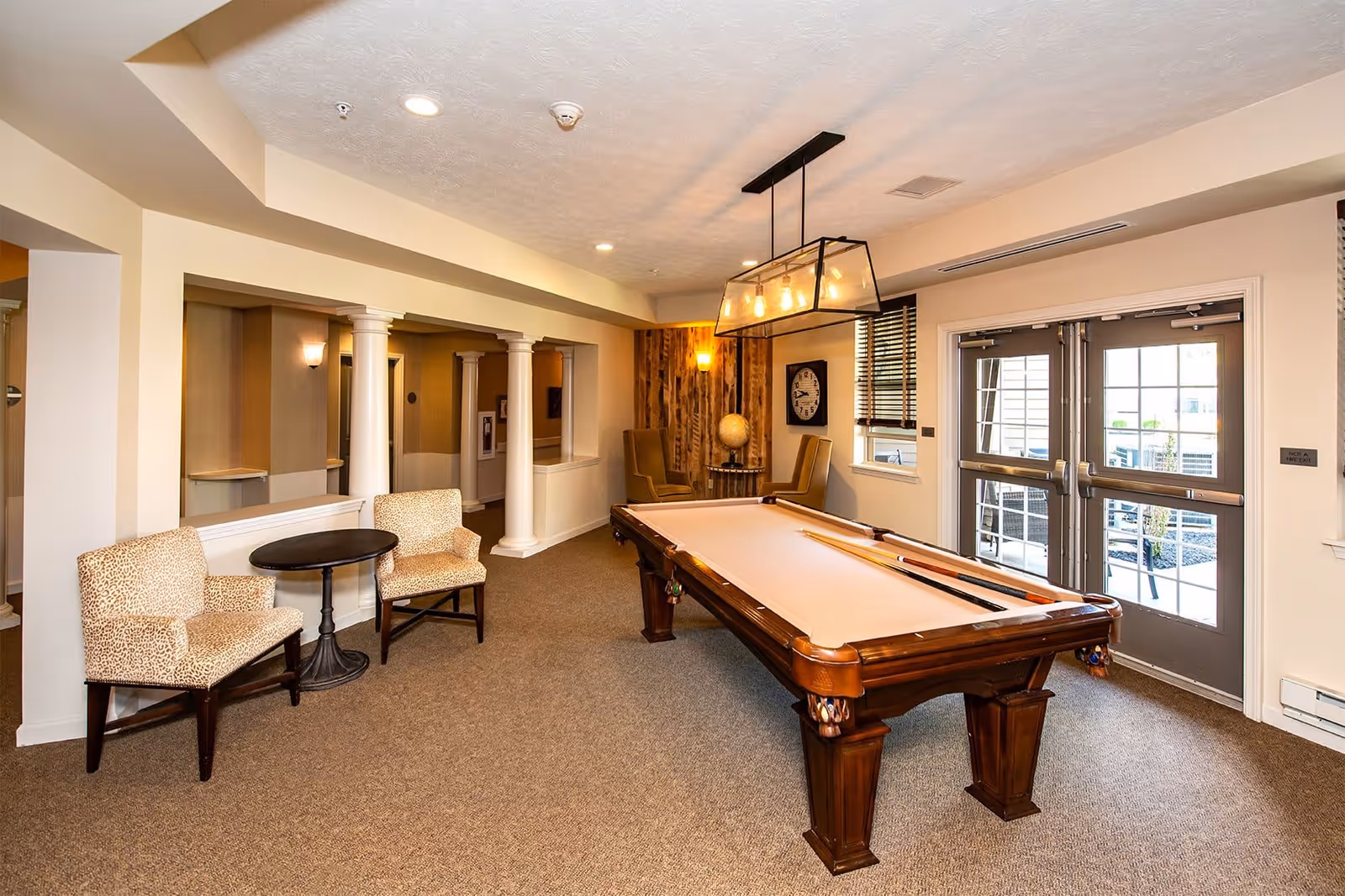 A well-lit recreational room featuring a pool table with cues on top, two patterned armchairs with a small round table between them, and a seating area with two high-back chairs and a globe on a side table. The room has beige walls, carpeted floor, white columns, and double glass doors leading outside.