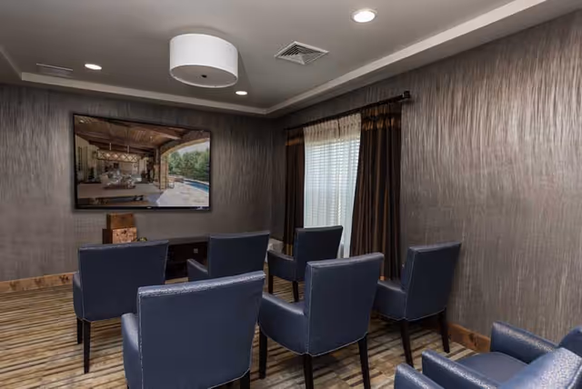 Small media room with rows of blue upholstered chairs facing a wall-mounted flat-screen TV and a curtained window.