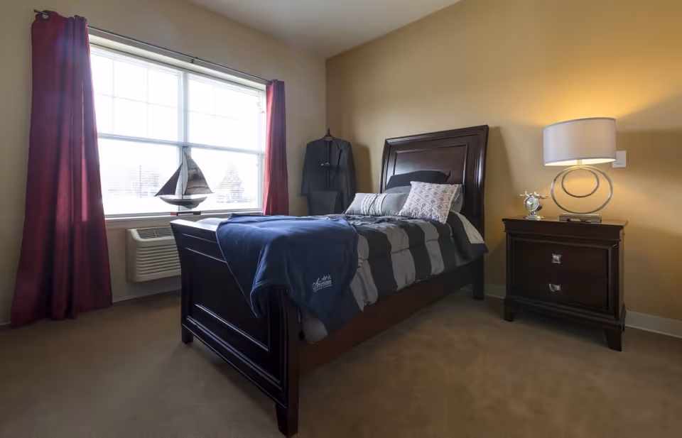 A cozy bedroom with a single dark wooden bed featuring gray and black striped bedding and multiple pillows. A navy blue blanket with 'All American Assisted Living' embroidered on it is draped over the foot of the bed. Next to the bed is a matching dark wooden nightstand with a modern lamp and a small decorative clock. A window with red curtains lets in natural light, and a decorative sailboat is placed on the windowsill. A suit jacket is hanging on a stand in the corner of the room.