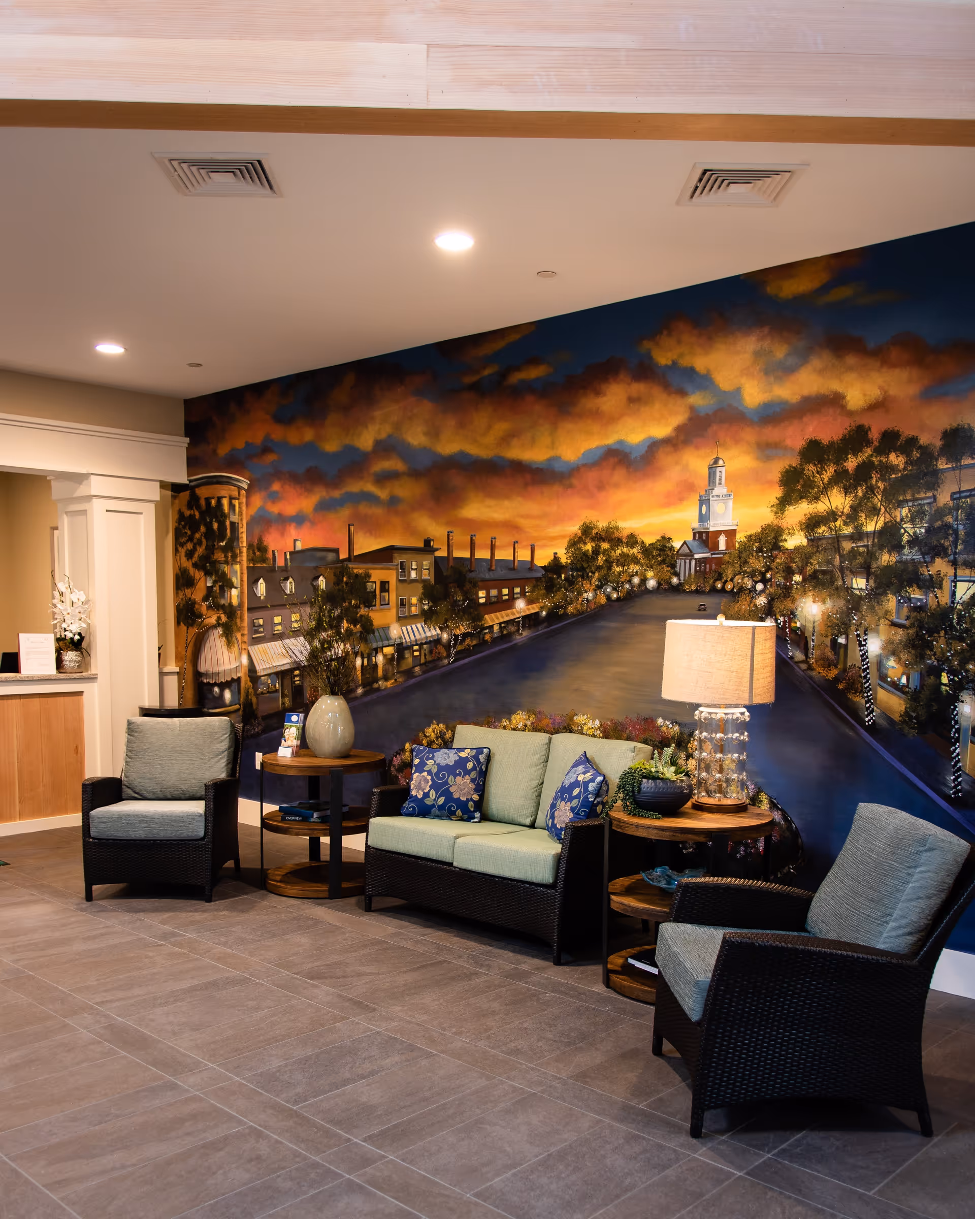 A cozy seating area with two armchairs and a loveseat featuring green cushions and blue floral pillows, arranged around two wooden side tables with decorative items and a lamp. Behind the seating is a large mural depicting a sunset sky over a town street with buildings and trees.