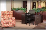 Interior lounge with a floral upholstered recliner, wooden desk and chair, potted plants, and a lamp.