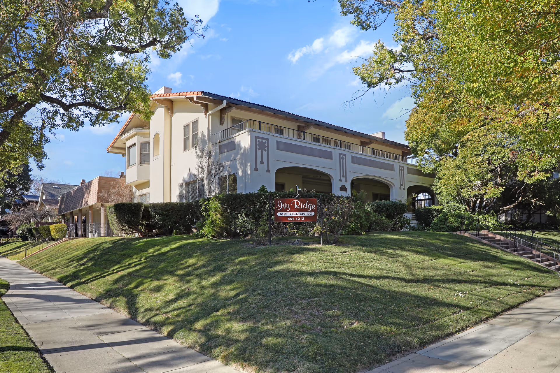 Exterior view of Ivy Ridge Assisted Living facility, a two-story building with a beige facade, decorative architectural details, and a red tile roof. The building is surrounded by green grass, bushes, and large trees under a blue sky with some clouds. A sidewalk runs along the front of the property, and a sign in front reads 'Ivy Ridge Assisted Living 451-1212'.