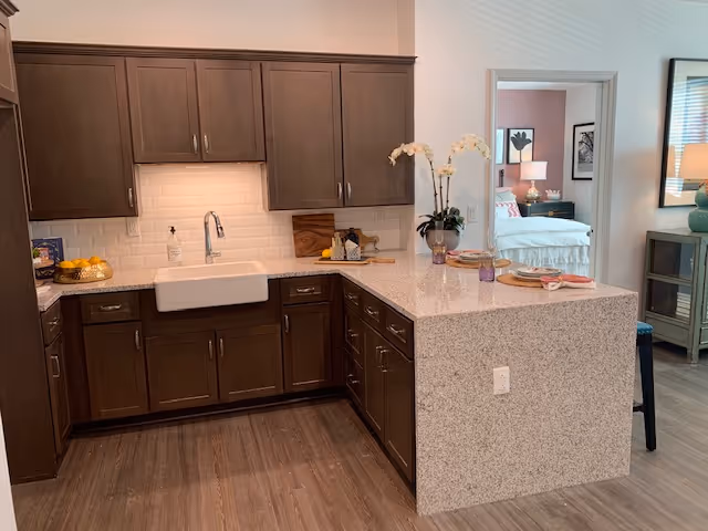 A modern kitchen with dark wooden cabinets, a white farmhouse sink, and a light-colored granite countertop that extends into a breakfast bar. The backsplash is made of white subway tiles. On the countertop, there are decorative items including a vase with white orchids and some kitchen essentials. In the background, there is a view into a bedroom with a bed, nightstand, and lamp.