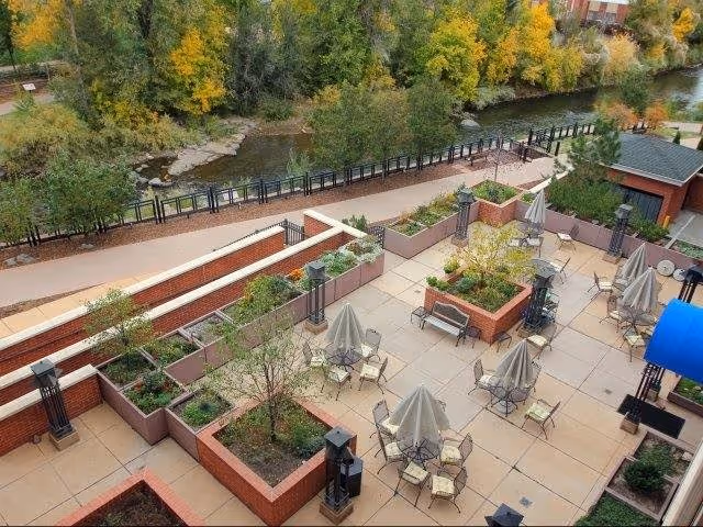 Aerial view of an outdoor patio area with several tables and chairs, each table shaded by a closed umbrella. The patio is surrounded by brick planters with small trees and plants. Beyond the patio, there is a walking path and a river bordered by trees with autumn foliage.