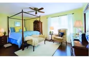 A bright and spacious bedroom featuring a four-poster bed with blue bedding, a cushioned bench at the foot of the bed, a beige armchair with a side table and lamp, a ceiling fan, and large windows with sheer white curtains allowing natural light to fill the room.