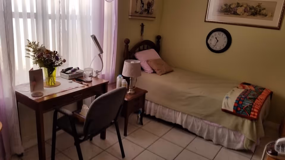 A small bedroom with a single bed covered with a beige blanket and colorful quilt at the foot. There are two pillows on the bed. Next to the bed is a wooden nightstand with a lamp and some bottles. A wooden desk with a chair is positioned near a window with sheer curtains, holding a vase of flowers, a lamp, a glass of water, and some other small items. A clock and framed pictures hang on the light-colored wall.