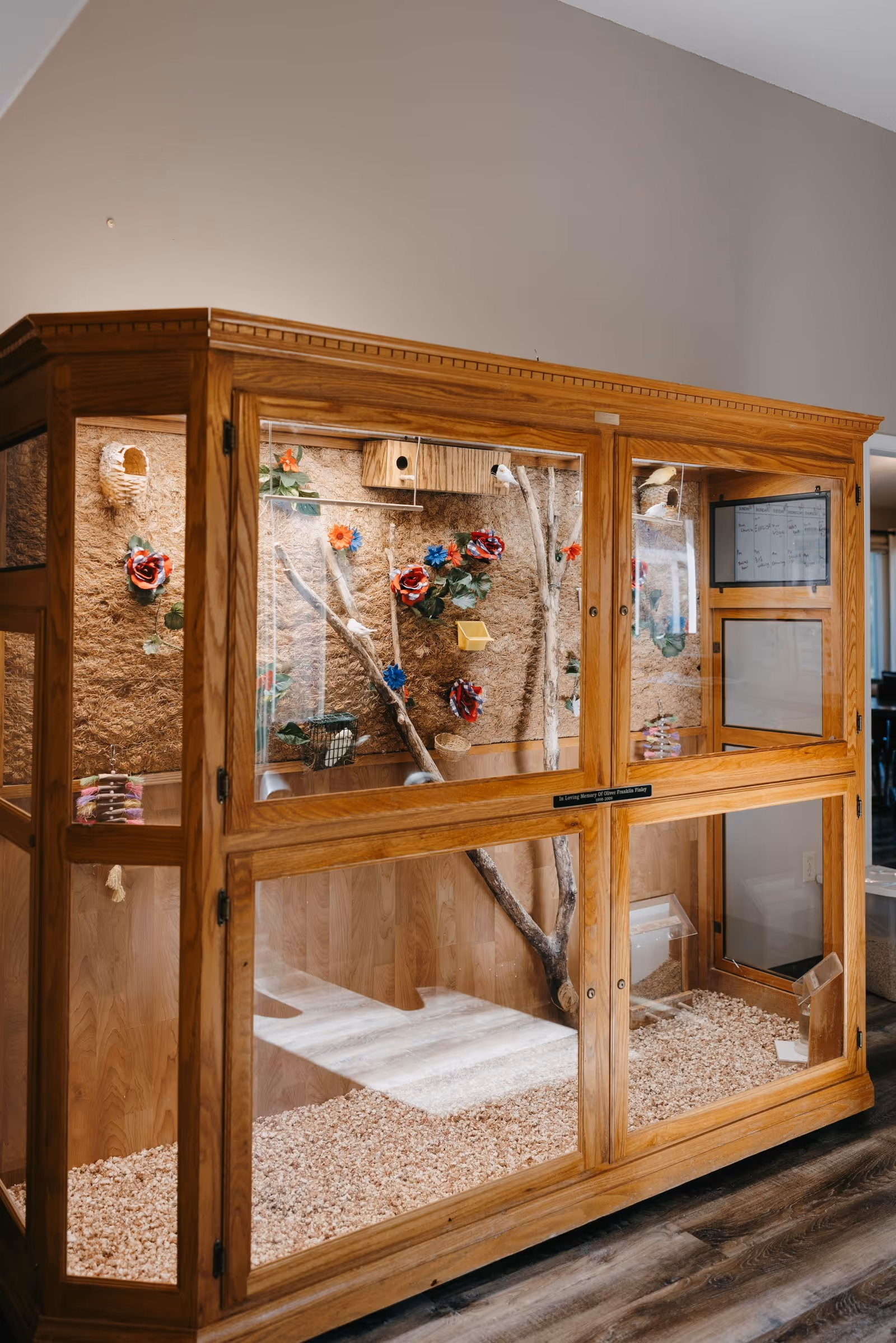A large wooden and glass enclosure designed as a habitat with branches, colorful artificial flowers, and small baskets inside. The enclosure is placed indoors on a wooden floor with a beige wall in the background.