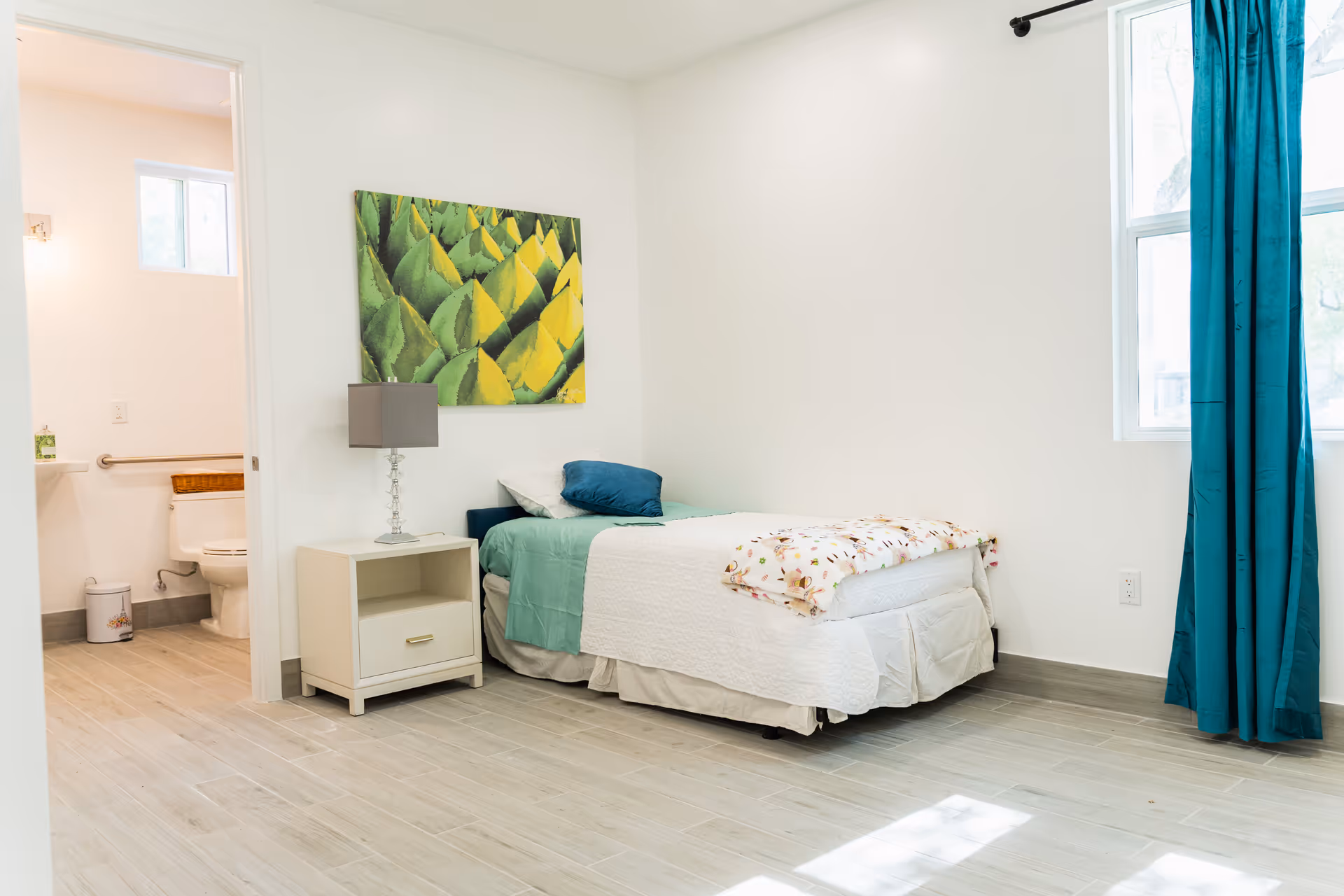 A bright, clean bedroom with a single bed made with white and green bedding, a small nightstand with a lamp, a colorful green and yellow abstract painting on the wall, and a window with teal curtains. An adjoining bathroom with a toilet and grab bar is visible through an open door.