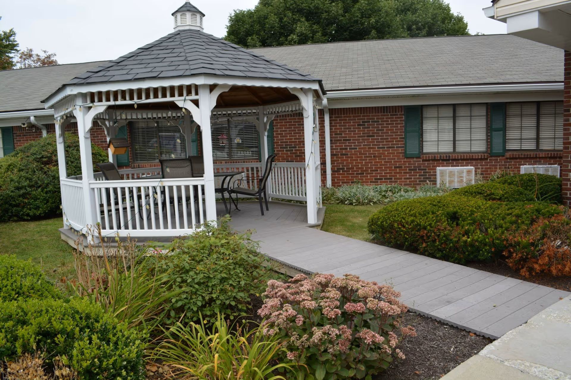 A white wooden gazebo with a gray shingled roof is situated in a garden area with various green shrubs and flowering plants. The gazebo contains outdoor chairs and a small table. Behind the gazebo is a brick building with green shutters and windows with blinds. A gray wooden pathway leads to the gazebo.