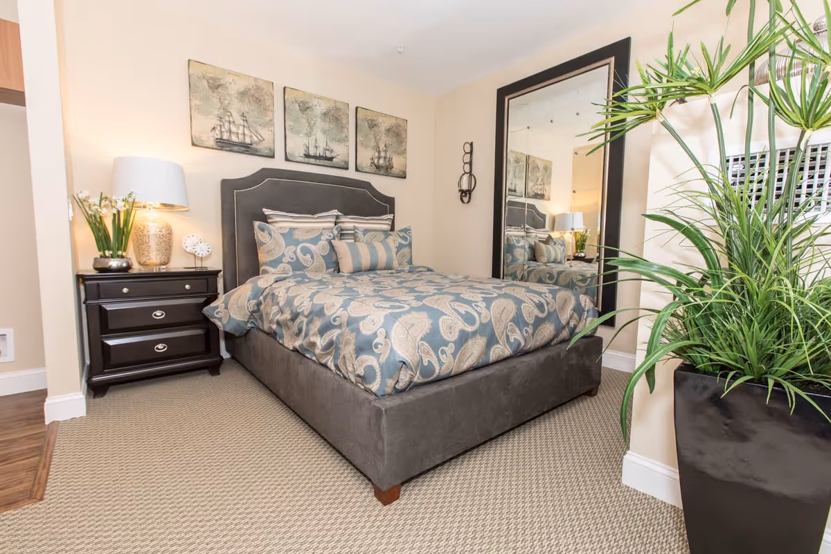 A neatly made bed with a gray upholstered headboard and blue paisley-patterned bedding in a bedroom. There is a black nightstand with a lamp, a small clock, and a vase with flowers on it. Above the bed are three framed pictures of old ships. A large mirror with a black frame is positioned against the wall to the right of the bed. A tall green plant in a black pot is in the foreground on the right side of the image.