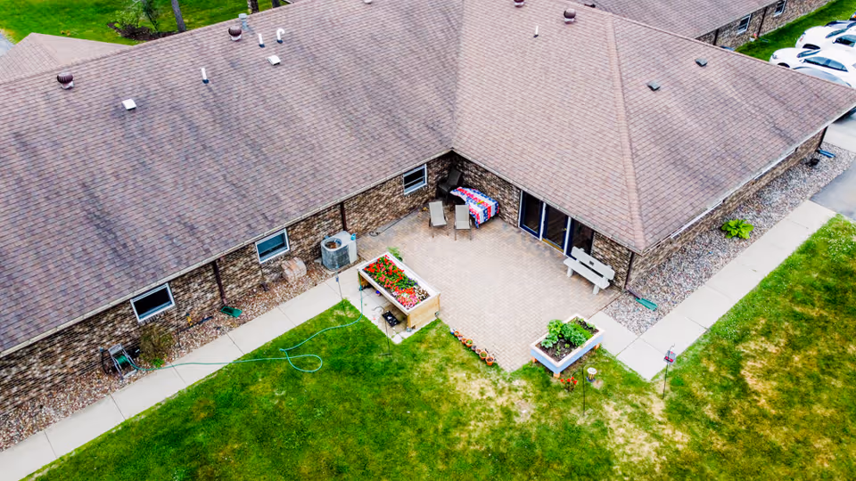 Aerial view of a single-story brick building with a brown shingled roof, a paved patio area with a table covered by a colorful cloth and two chairs, two raised garden beds with flowers and plants, a bench, and surrounding green lawn with sidewalks.