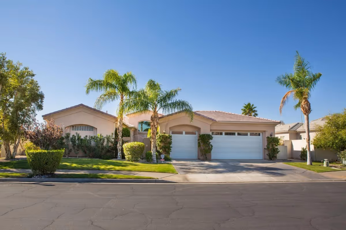 Front exterior view of a single-story residential building with a three-car garage, palm trees, and well-maintained landscaping under a clear blue sky.