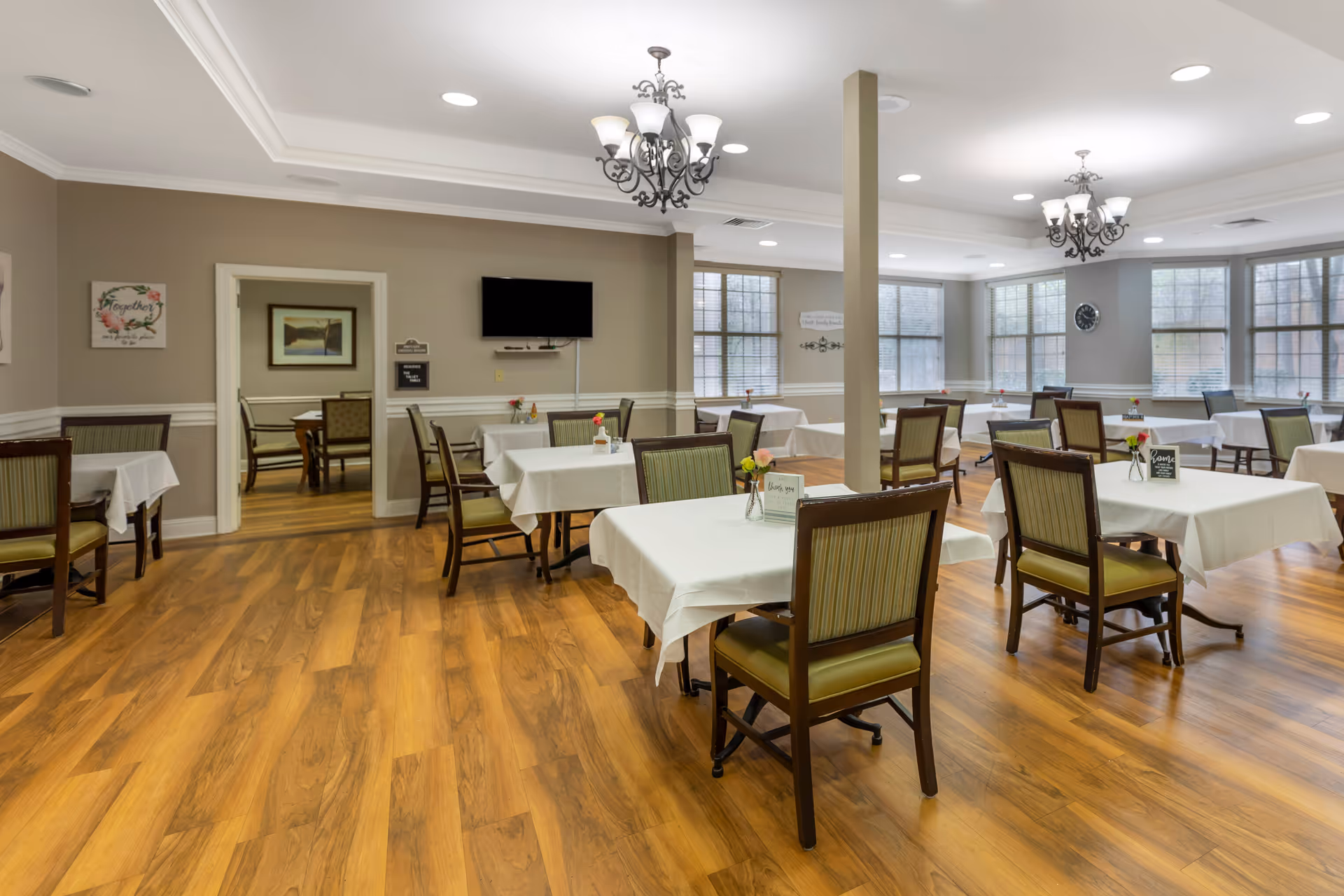 A dining room in a senior living facility with multiple tables covered in white tablecloths, each surrounded by wooden chairs with green cushions. The room has wood flooring, large windows letting in natural light, two chandeliers, and a wall-mounted TV. There are small flower vases and table signs on each table.