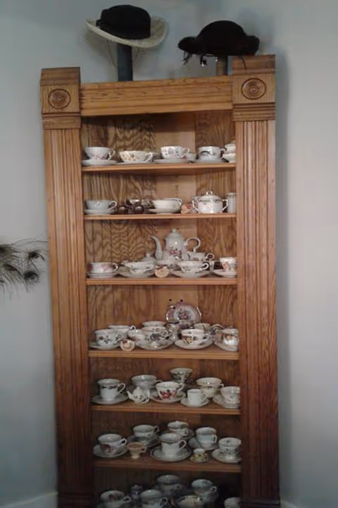 Tall wooden display shelf filled with china teacups, saucers and teapots, with hats resting on top against a white wall.