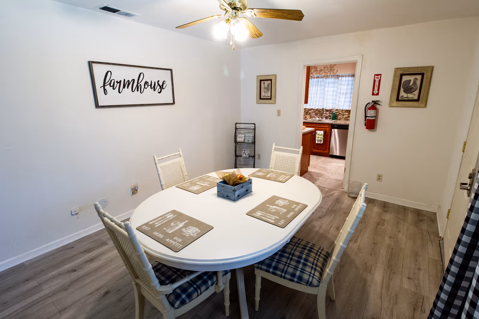 A dining room with a white oval table set with four chairs that have blue plaid cushions. The table has four placemats and a centerpiece with fruit. On the wall is a framed sign that reads 'farmhouse'. The room has light wood flooring and white walls. A doorway leads to a kitchen with wooden cabinets, a dishwasher, and a window with a curtain. There is a fire extinguisher mounted on the wall near the doorway.