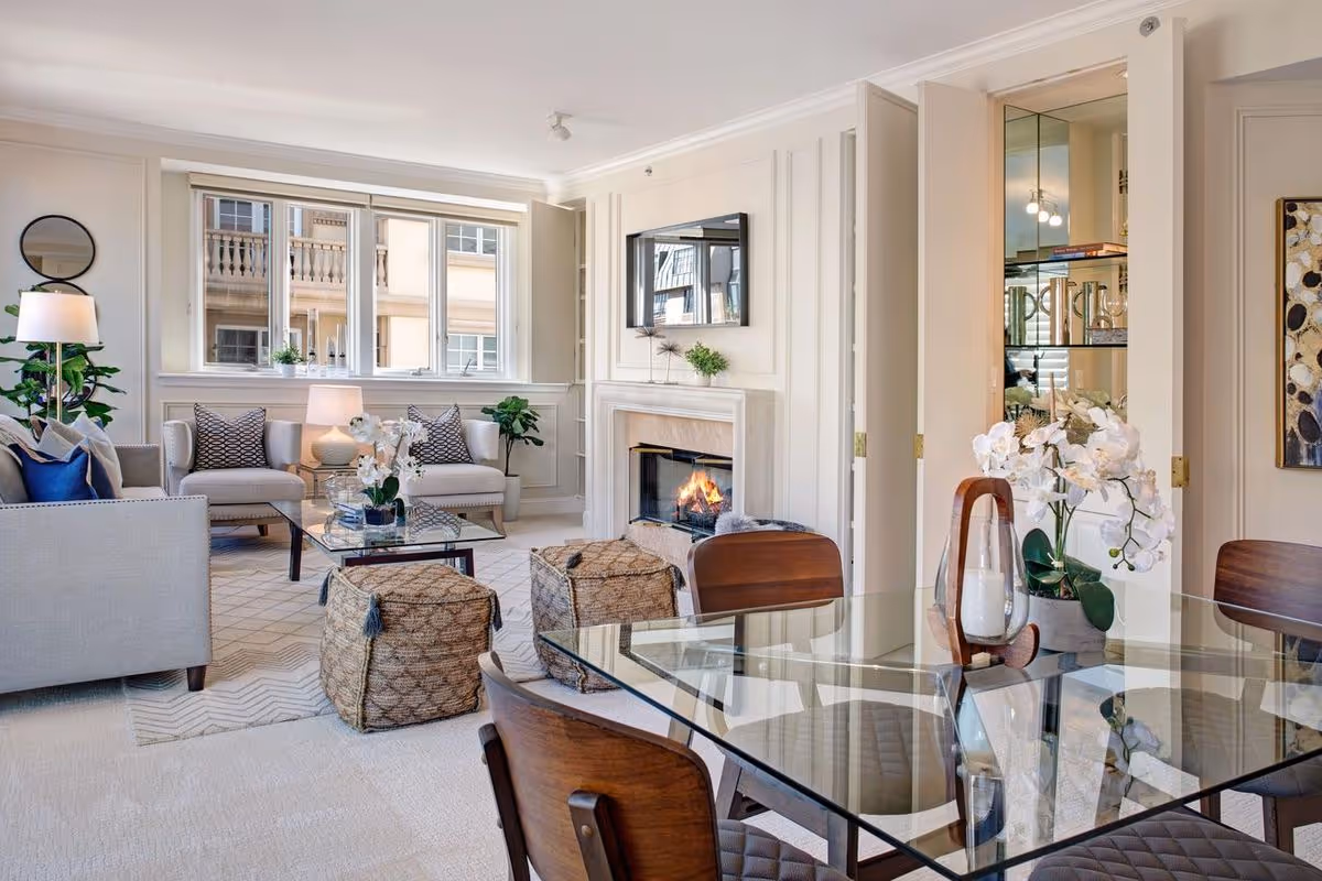 Bright and elegant living room with a glass dining table and wooden chairs in the foreground, a cozy seating area with two armchairs, a sofa, and two woven ottomans around a glass coffee table, a lit fireplace with a mounted TV above it, large windows letting in natural light, and decorative plants and artwork.