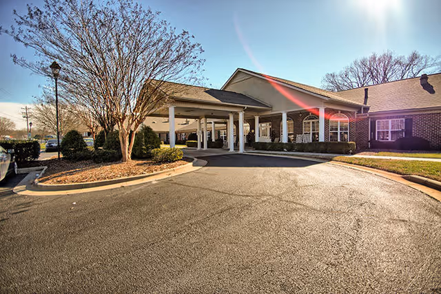 Exterior view of a senior living facility with a covered entrance supported by white columns, surrounded by landscaped bushes and trees under a clear blue sky.