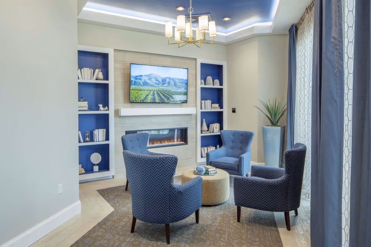 A cozy living room area with four blue patterned armchairs arranged around a round beige ottoman. The room features a modern electric fireplace set into a tiled wall with a flat-screen TV mounted above it. Built-in shelves with books and decorative items flank the fireplace. A tall plant in a blue pot stands near large windows with sheer and blue curtains. The ceiling has recessed lighting and a modern chandelier.