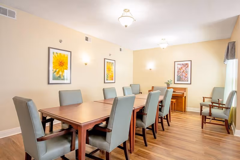 A well-lit dining room with a long wooden table surrounded by eight padded chairs with armrests. The walls are painted light beige and decorated with framed floral artwork. There is a wooden piano against the far wall with a small plant on top, and two additional chairs are placed near a window with curtains. The floor is a light wood laminate.