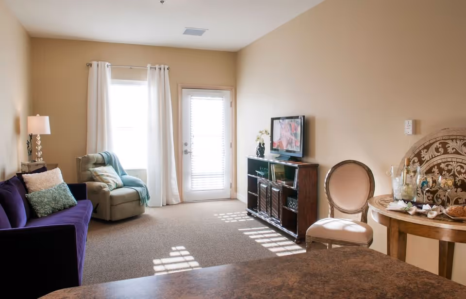 A cozy living room with beige walls and carpeted floor. The room features a purple sofa with decorative pillows, a green armchair with a throw blanket, a wooden TV stand with a flat-screen TV, and a round wooden table with a chair. Sunlight streams through a window with white curtains and a glass door with blinds.
