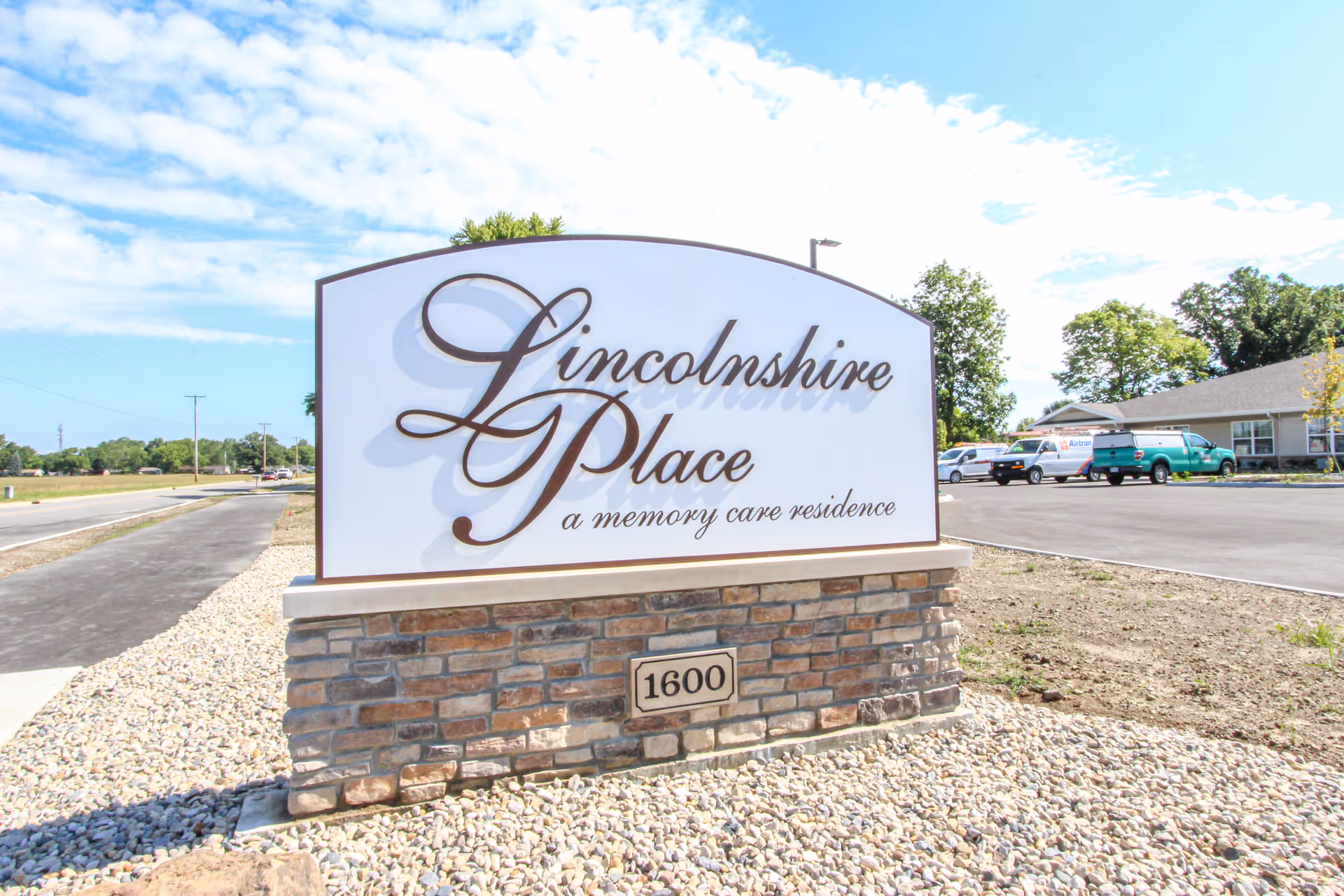 Outdoor view of a large sign for Lincolnshire Place, a memory care residence, with a brick base and white top featuring elegant script. The sign is located near a paved road and parking lot with some vehicles and a building in the background under a partly cloudy sky.