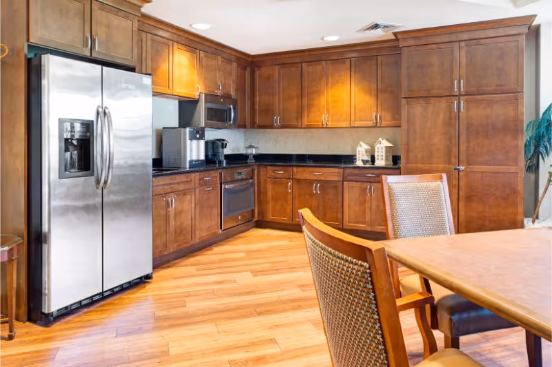 A modern kitchen area with wooden cabinets, a stainless steel refrigerator, built-in microwave, oven, coffee maker, and a dining table with chairs. The floor is wooden and the space is well-lit with recessed ceiling lights.