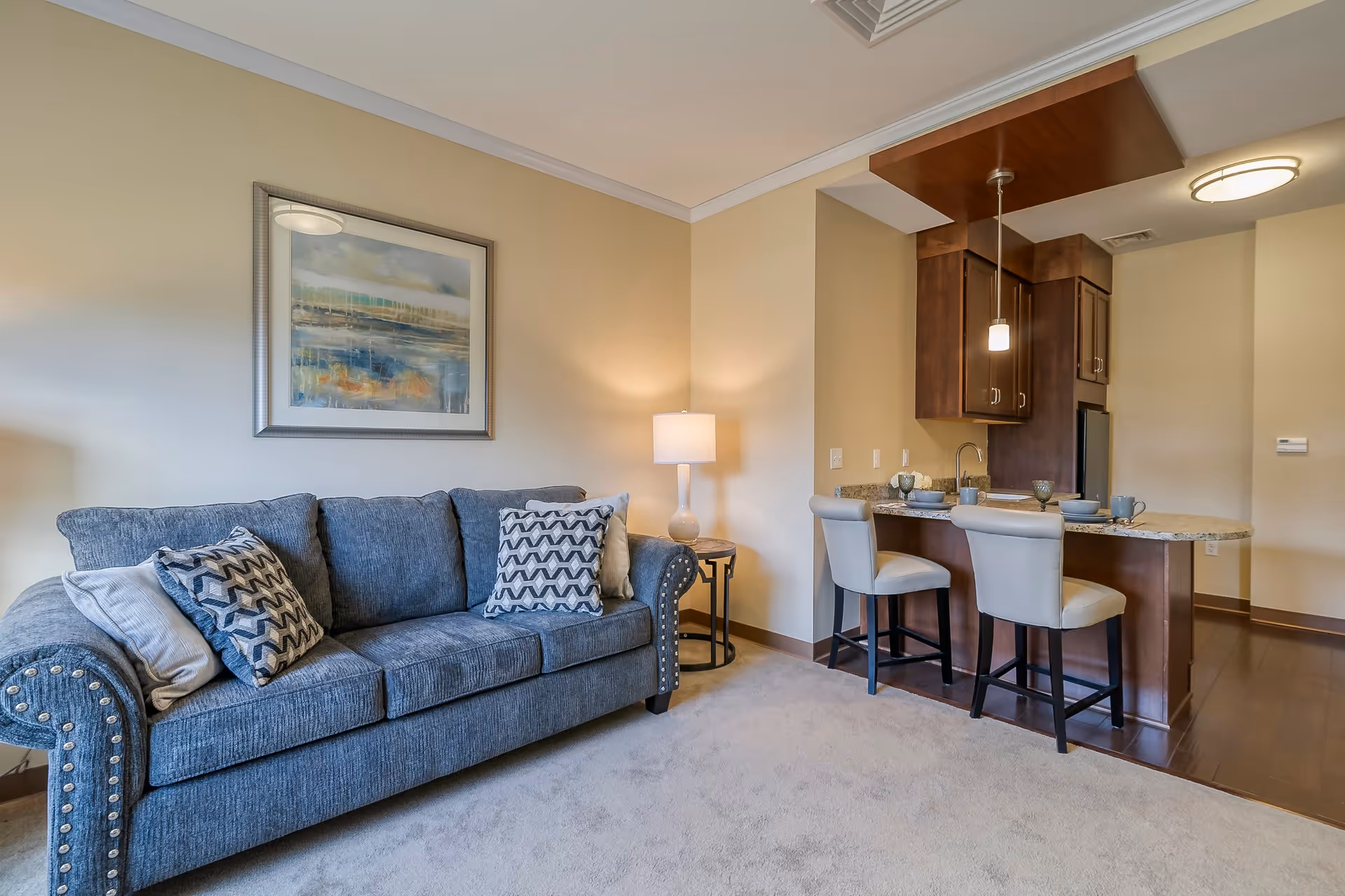 A cozy living area featuring a blue upholstered sofa with patterned cushions, a side table with a lamp, and a framed abstract painting on the wall. Adjacent to the living area is a small kitchen with dark wood cabinets, a granite countertop breakfast bar with two cushioned stools, and pendant lighting above.
