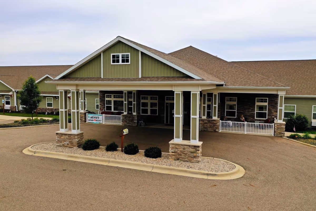 Front exterior view of a single-story senior living facility building with a covered entrance supported by columns with stone bases. The building has green siding with stone accents and multiple windows. There is a small landscaped area with bushes and a mailbox near the entrance. A sign near the entrance reads 'HEROES WORK HERE!'.