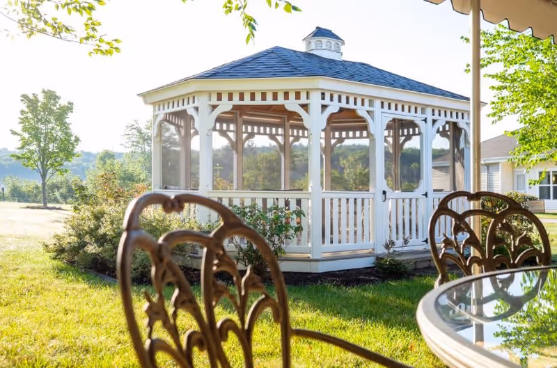 A white wooden gazebo with a blue roof situated on a grassy lawn surrounded by trees and bushes, with a glass-top table and ornate metal chairs in the foreground.