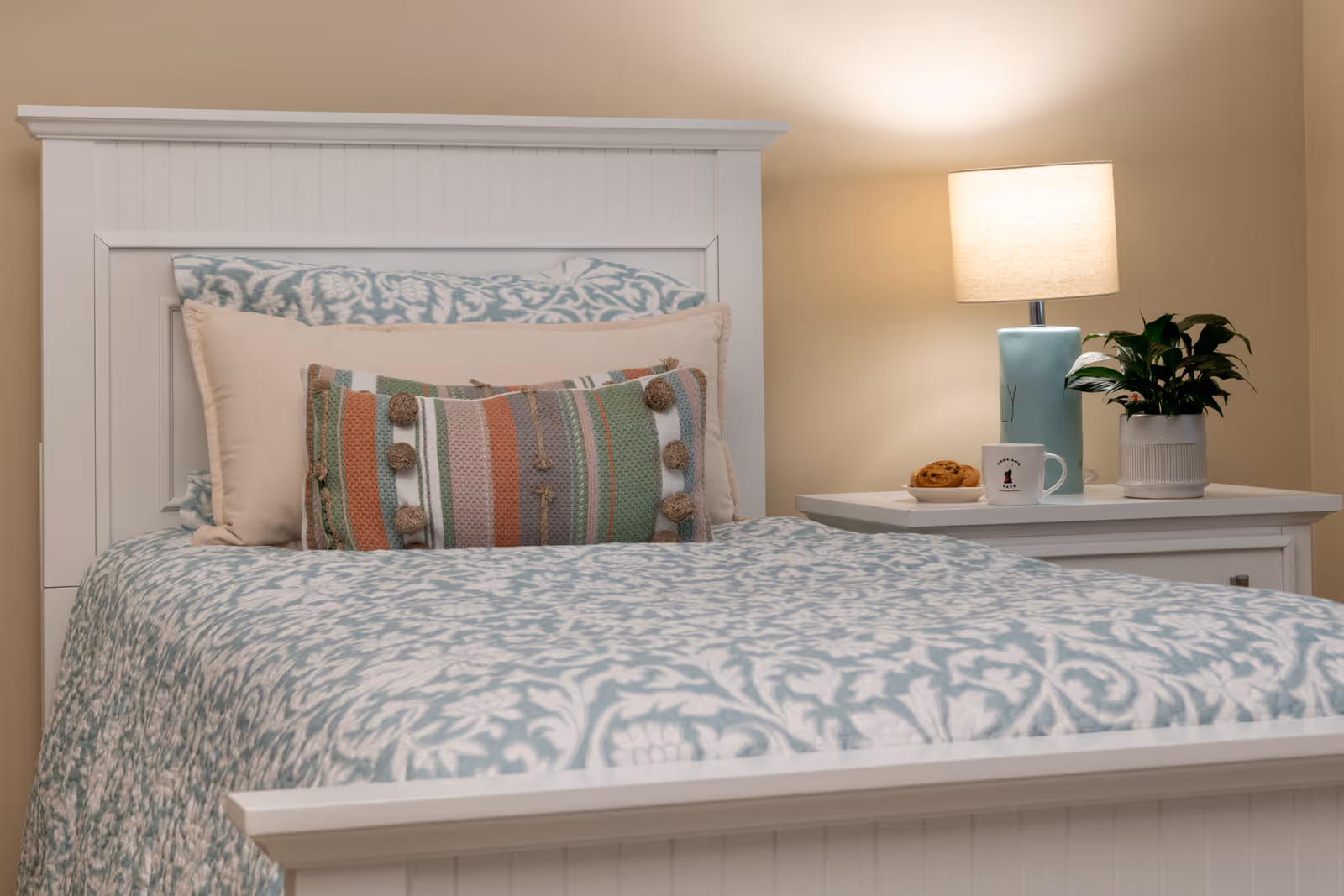 A neatly made bed with a white wooden headboard and footboard, covered with a blue and white patterned quilt. There are three pillows arranged at the head of the bed, including a decorative pillow with multicolored stripes and pom-poms. Next to the bed is a white nightstand with a blue lamp, a small potted plant, a coffee mug, and a plate with a cookie. The wall behind the bed is painted a light beige color.