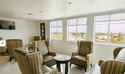 A bright common area with several striped armchairs arranged around small round tables near large windows that offer a view of the outdoors. The room has a clean, modern look with white walls and a mounted flat-screen TV.