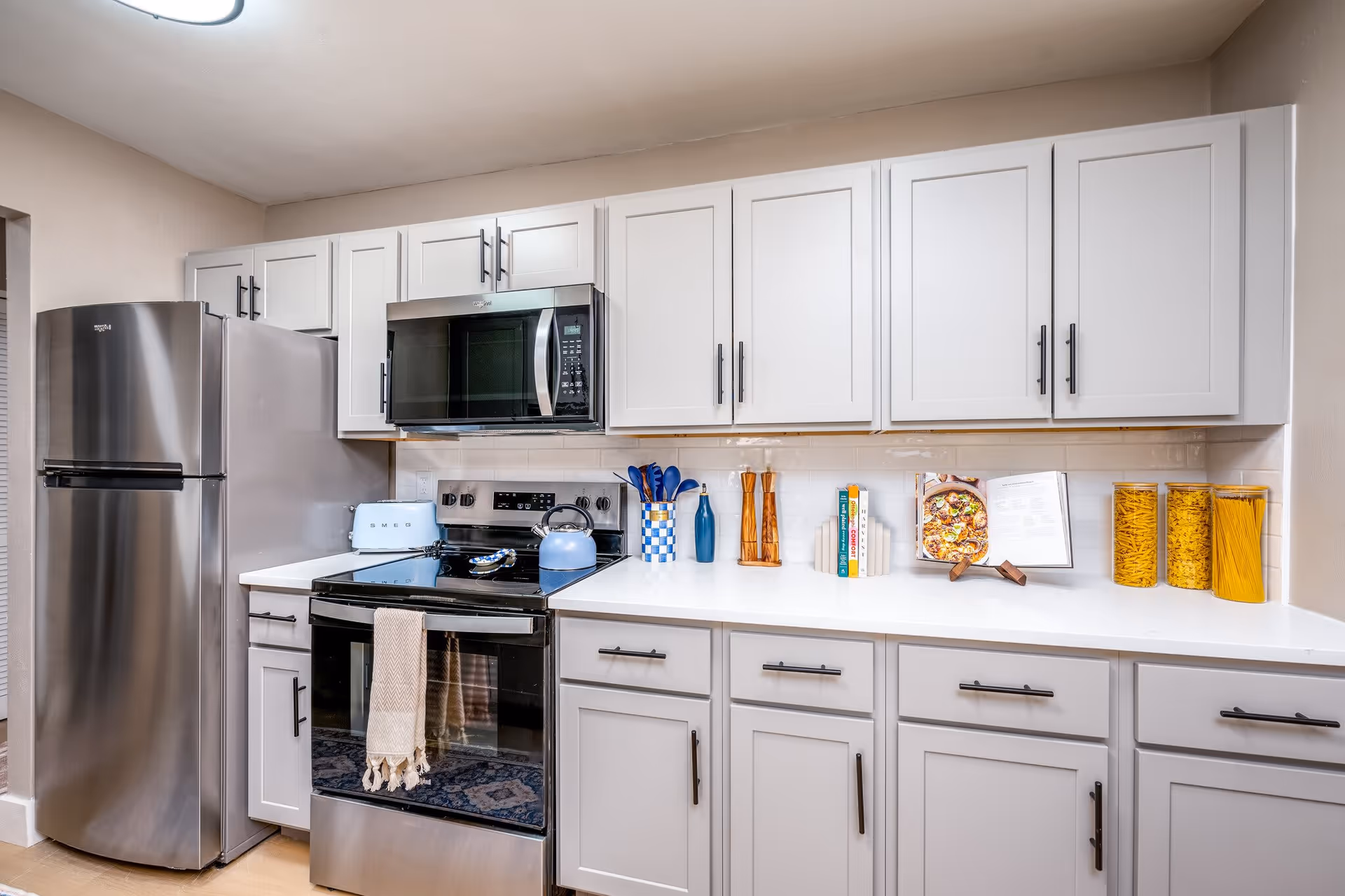 A modern kitchen with white cabinets and black handles, a stainless steel refrigerator, a black stove with a beige towel hanging on the handle, and a black microwave above the stove. The countertop has a blue toaster, a blue kettle, a utensil holder with blue utensils, two wooden pepper mills, a few cookbooks, and three clear containers filled with pasta.